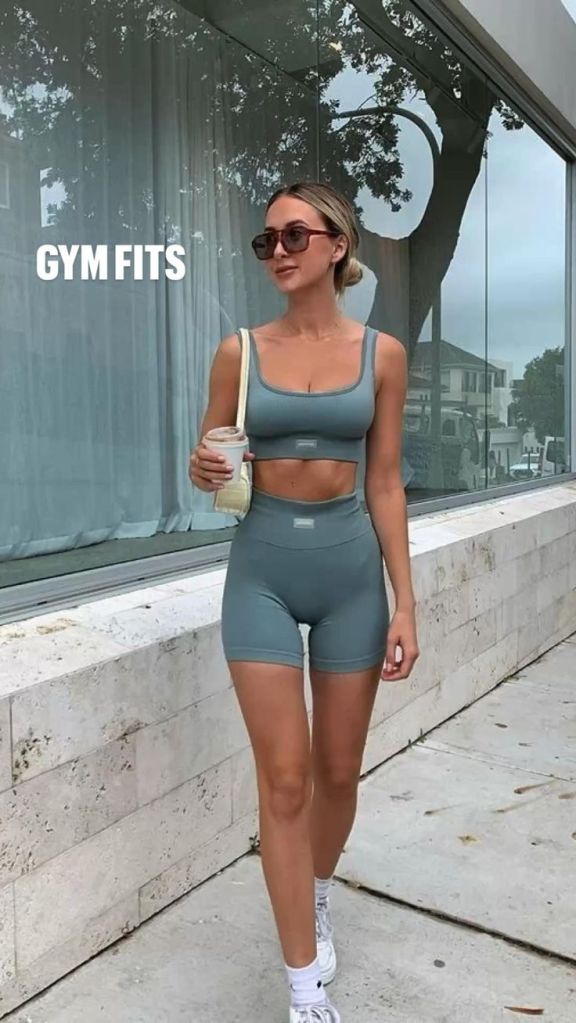A woman walking in sporty gym attire, wearing a light blue crop top and matching shorts, holding a beverage in her right hand, with sunglasses on, against a backdrop of a modern building.